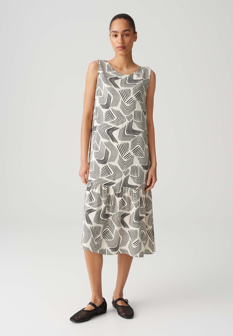Sleeveless midi dress in cream with black geometric wave pattern, featuring a ruffled hem. Worn with black mesh flats. Smooth fabric texture.