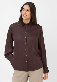 Blusa lunga a maniche con colletto classico, di colore marrone scuro, con chiusura a bottoni e una leggera lucentezza; presenta polsini arricciati e una vestibilità morbida.