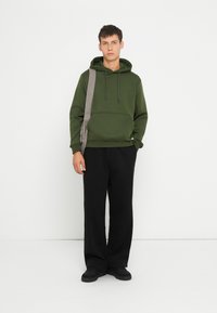 Felpa verde con tasca frontale, cappuccio con lacci e polsini a coste, abbinata a pantaloni neri larghi e scarpe nere.