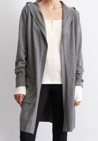 Grå lång kofta i mjukt tyg, med huva, öppen front och sidofickor. Över en vit ribbad topp och svarta byxor.