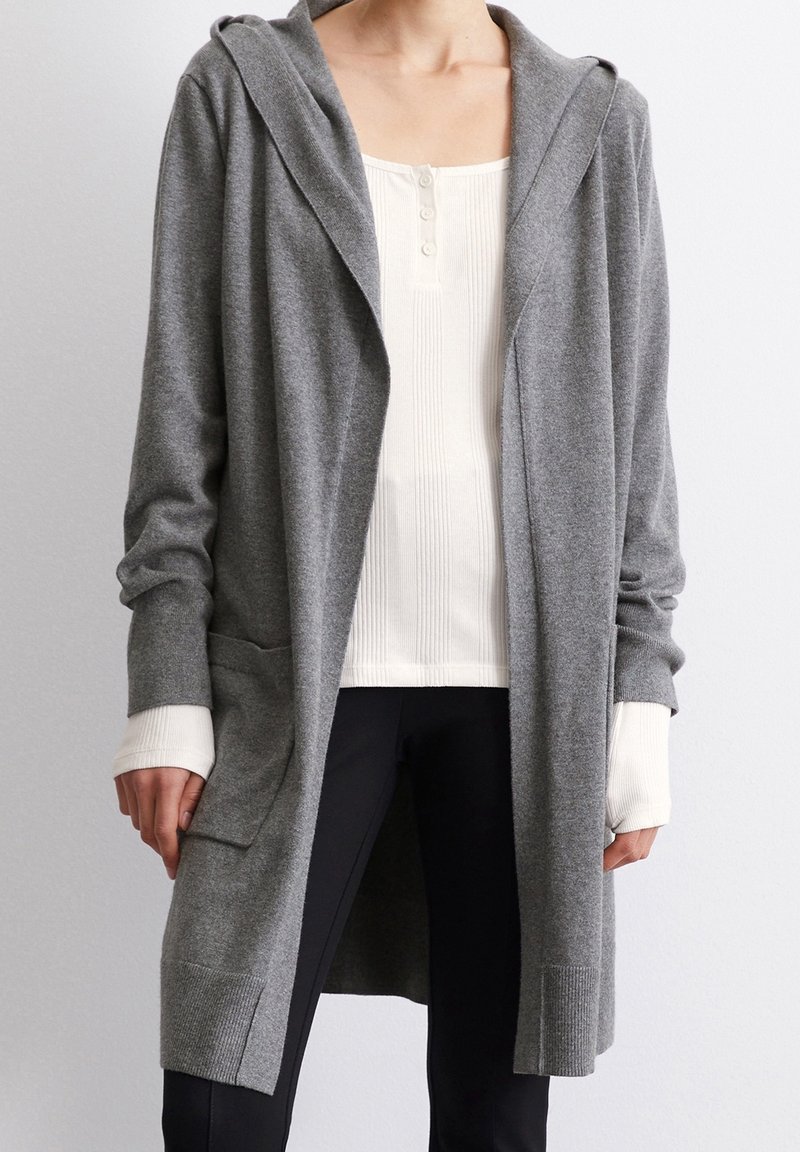 Grå lång kofta i mjukt tyg, med huva, öppen front och sidofickor. Över en vit ribbad topp och svarta byxor.