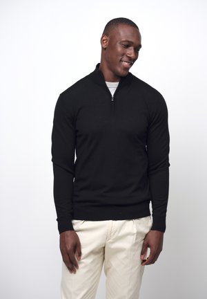 Black long-sleeve zip-up sweater with a collared neck, ribbed cuffs, and hem, paired with light beige trousers. Smooth texture, classic fit.