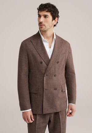 Homme portant un costume marron croisé à revers crantés et une chemise blanche, regardant légèrement sur le côté sur un fond neutre.