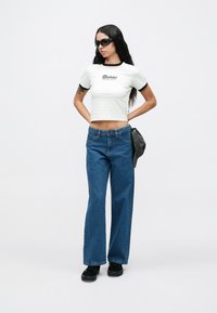 Γυναίκα που φοράει λευκό crop t-shirt με λογότυπο, μπλε τζιν ψηλόμεσο με φαρδιά πόδια, μαύρα αθλητικά παπούτσια, γυαλιά ηλίου και κρατάει ένα γκρι καπέλο πίσω από την πλάτη της.