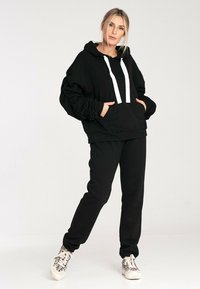 Czarna bluza z kapturem z białymi sznurkami i kieszenią z przodu, w połączeniu z czarnymi spodniami dresowymi. Oba elementy wykonane są z miękkiego, strukturalnego materiału.