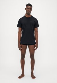 Svart kortärmad t-shirt med rund halsringning, med texturerade detaljer vid axlarna och logotypdetalj på bröstet. Bärs med svarta shorts.