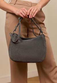 Graue Wildleder-Handtasche mit einer geschwungenen Form, ausgestattet mit verstellbaren Trägern und silbernen Hardware-Details, gehalten von Händen.