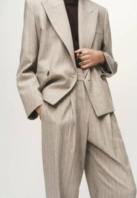 Blazer beige à rayures avec de larges revers et une coupe ajustée, associé à un pantalon plissé taille haute. Comprend des boutons dorés et des bagues superposées.