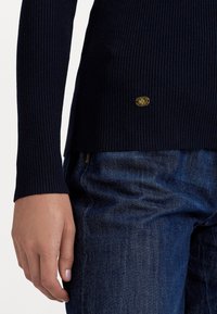 Suéter riscado azul-marinho de mangas compridas, com um pequeno emblema de logotipo dourado e detalhe na barra, combinado com jeans de denim escuro.