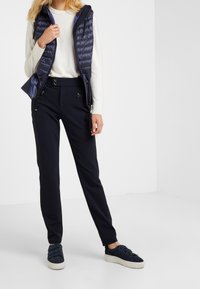 Gilet en duvet marine avec poches zippées sur un pull blanc, pantalon marine assorti et mocassins en daim bleu avec une semelle blanche.