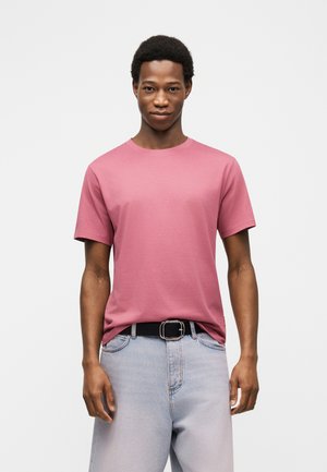 Jeune homme portant un t-shirt rose uni à manches courtes, rentré dans un jean bleu clair avec une ceinture noire, debout devant un fond uni.