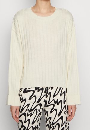 Maglione a coste di colore crema, con scollatura a girocollo e maniche oversize, abbinato a pantaloni a motivi bianco e nero.