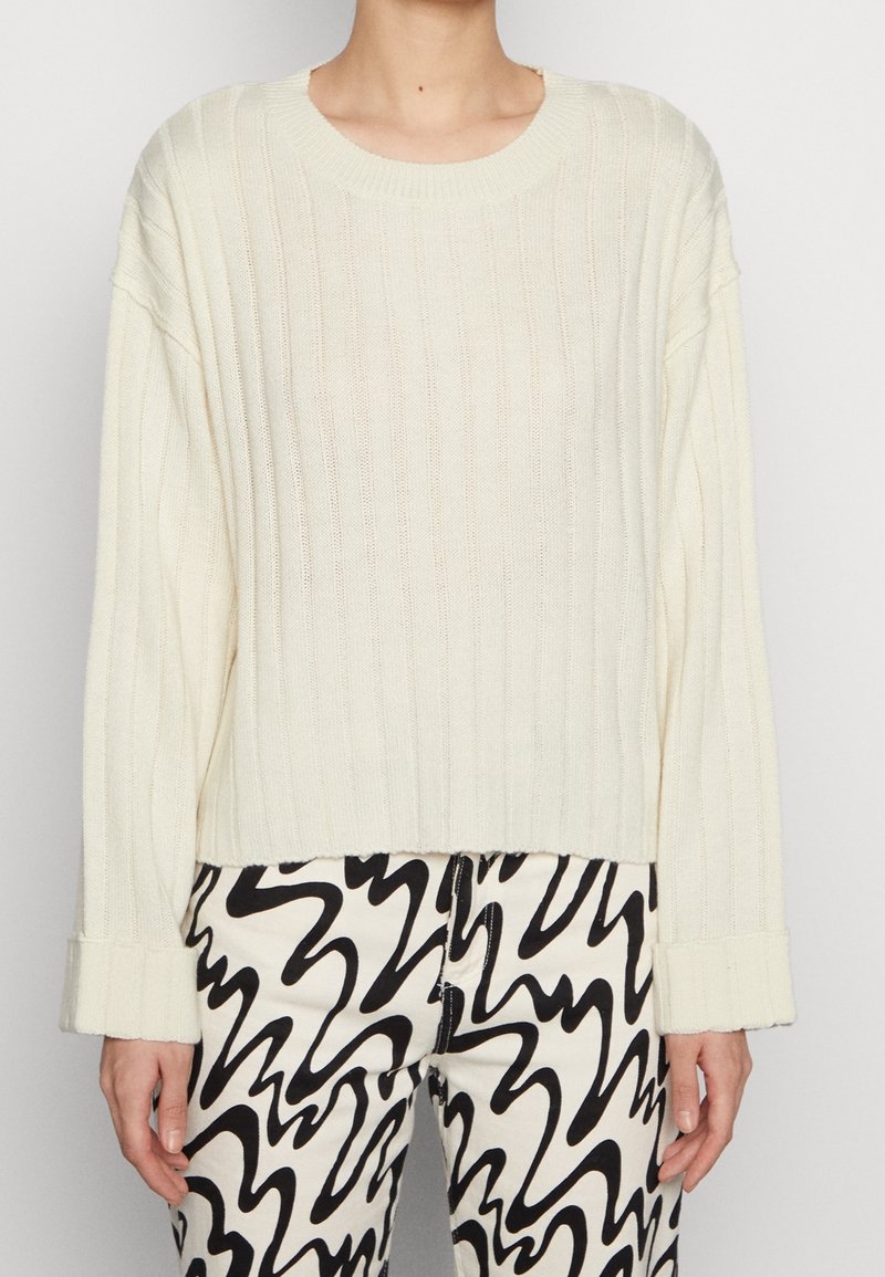 Maglione a coste di colore crema, con scollatura a girocollo e maniche oversize, abbinato a pantaloni a motivi bianco e nero.