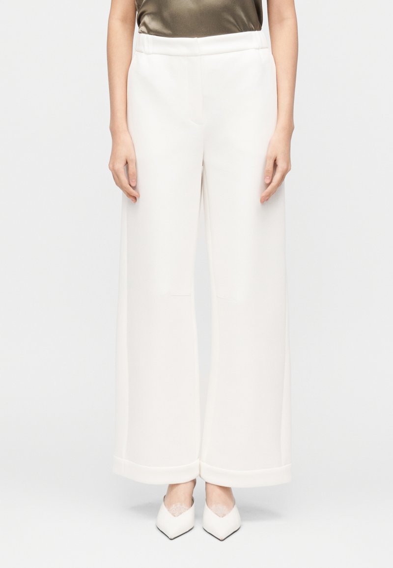 Calças brancas de perna larga feitas de tecido liso, com cintura alta e bainhas viradas. Combinadas com sapatos brancos pontiagudos.