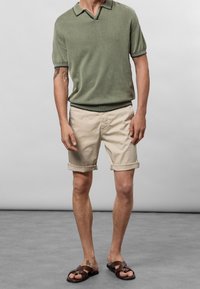 Polo en tricot vert avec un col côtelé, shorts roulés beige et sandales en cuir marron. Tenue d'été décontractée avec une coupe ample.