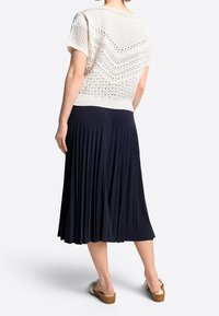 Hvid strikket top med korte ærmer og åben mønster, kombineret med en tætsiddende, plisseret marineblå nederdel, båret med beige sandaler.