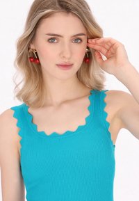 Turquoise geribbelde tanktop met golvende randen, gecombineerd met rode kersenvormige oorbellen. Zachte textuur, aansluitend ontwerp en levendige kleuren.