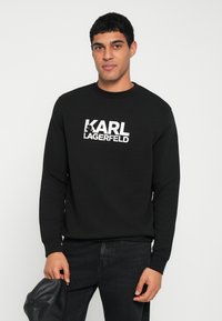 Felpa nera con maniche lunghe e scollo tondo, caratterizzata da un logo bianco a contrasto "KARL LAGERFELD" sul davanti.