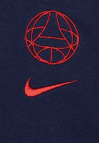 La tela azul marino presenta logotipos bordados en rojo: un diseño de globo sobre el swoosh de Nike. Textura suave con una apariencia limpia y minimalista.