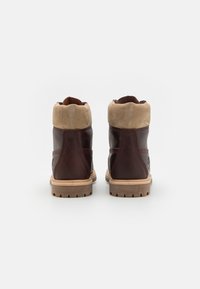 Botas de couro castanho com um colarinho em camurça bege, biqueira redonda e sola de borracha robusta, apresentando detalhes em costura e um logo na lateral.