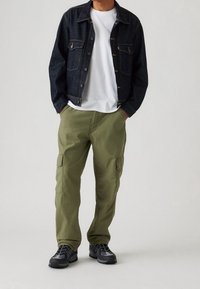 Donkere spijkerjacket met borstzakken, witte T-shirt, olijfgroene cargobroek met zijzakken, en zwarte sportschoenen met grijze accenten.