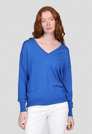 Pull bleu en V en tissu doux, avec des manches longues et une coupe décontractée. Accompagné d'un pantalon blanc, sur un fond gris.