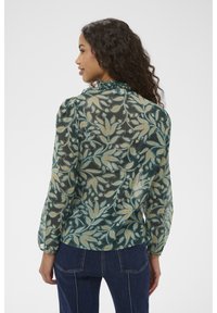 Mujer con cabello rizado que lleva una blusa transparente con estampado floral en verde, azul y marrón claro, combinada con vaqueros azul oscuro, de espaldas y mirando hacia la izquierda.
