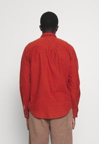 Rood overhemd met lange mouwen en een geknoopte kraag, gemaakt van een gestructureerde stof met een afgeronde zoom, gezien vanaf de achterkant.