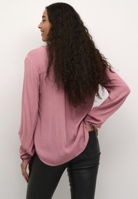Langärmlige rosa Bluse aus strukturiertem Stoff, mit knöpfbaren Manschetten und abgerundetem Saum, kombiniert mit eng anliegenden schwarzen Lederhosen.