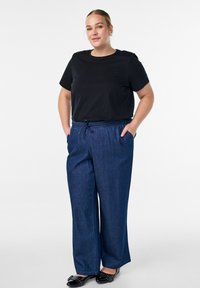 Moteris stovi su rankomis kišenėse, vilkėdama juodą marškinėlį, mėlynas plačias kelnes ir juodus odinius batus su studelėmis, priešais vienspalvį baltą foną.