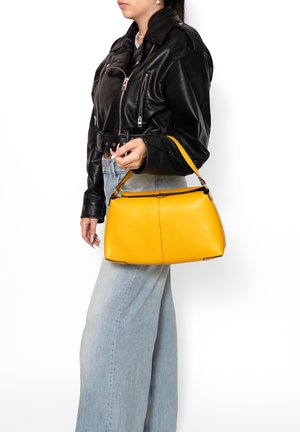 Donna che indossa una giacca di pelle nera e jeans azzurri chiari, che tiene una borsa gialla brillante per il manico al suo fianco.