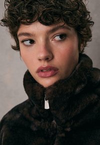 Brun kunstpelsjakke med høj krave og sølv lynlåsdetalje. Hudtonen er neutral med blød makeup og naturligt udseende krøller.