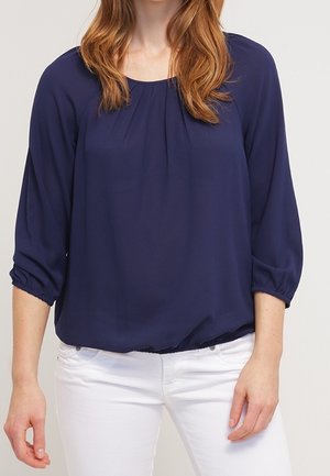 Femme portant un chemisier bleu marine ample à manches trois-quarts et un pantalon blanc, debout devant un fond uni.