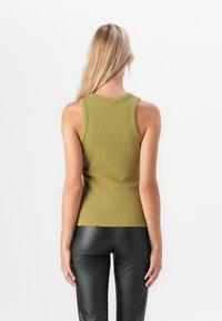 Kvinna med långt blont hår iförd en ärmlös ribbad topp i olivgrön färg och svarta läderbyxor, stående vänd bort från kameran mot en vit bakgrund.