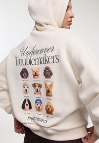 Felpa con cappuccio color crema con diverse illustrazioni di cani sul retro, con la scritta "Undercover Troublemakers" e una didascalia "Puppy Outlaws".