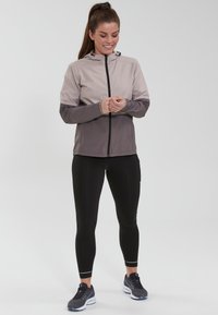 Lettet, to-farvet hættejakke i beige og mørkegrå, med lynlås foran og elastiske manchetter, parret med sorte leggings og grå sneakers.