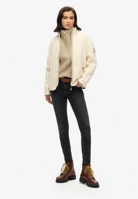 Witte fleece zip-up jas over een gestructureerde beige trui, gecombineerd met zwarte jeans en bruin veterschoenen. Minimalistisch ontwerp, functionele details.