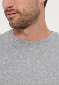 Άντρας με γένια που φοράει ανοιχτογκρι t-shirt με στρογγυλή λαιμόκοψη, κοιτάζοντας ελαφρώς αριστερά, με κοντινό πλάνο στο κάτω μέρος του προσώπου και του λαιμού του.