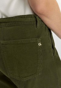 Primer plano de una persona que lleva pantalones de pana verde oliva con un bolsillo trasero cuadrado y una pequeña etiqueta metálica dorada.