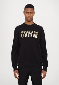 Felpa nera realizzata in tessuto dalla trama morbida, con la scritta "VERSACE JEANS COUTURE" in lettere dorate in grassetto sulla parte anteriore.