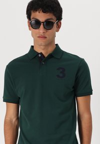 Tmavozelená polo košela vyrobená z pevného materiálu, s trojitému gombíkmi, námorníckym lemovaním goliera a logom s vyšitým číslom 3 na hrudi.
