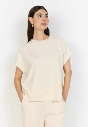 Frau mit langen dunklen Haaren, die ein lockeres, cremefarbenes Kurzarmshirt und passende Hosen trägt, Hände in den Taschen, schaut nach oben und rechts.