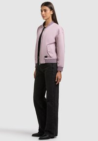 Femme debout de profil portant une veste bomber violet clair, un haut noir, un jean large noir et des bottines noires.