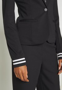 Blazer noir avec un design ajusté, fermeture à un bouton et poches latérales. Les poignets présentent des rayures blanches pour le contraste. Texture lisse.