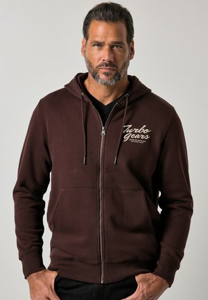 Mann mit kurzen Haaren und Bart, der einen dunkelbraunen Kapuzenpullover mit Reißverschluss und dem Text "Turbo Gears" auf der Brust trägt, steht mit den Händen in den Taschen.