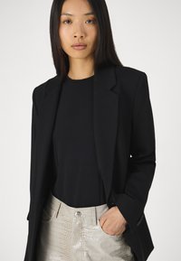 Black blazer with a structured design and notched lapels layered over a black top, paired with glossy, light beige crocodile-textured pants.