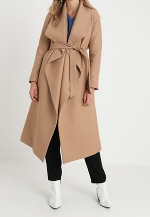 Femme portant un long manteau camel avec un large col et une ceinture, pantalon noir et bottines blanches sur un fond blanc uni.