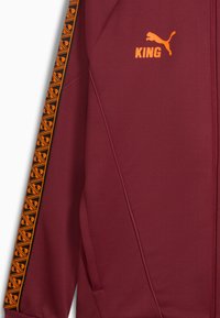 Maronjakke med orange detaljer; har et sort og orange mønstret stribe, "KING" logo og en lynlås foran.