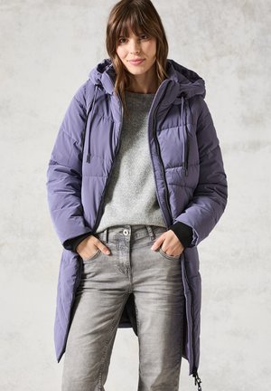 Lilla polstret lang frakke med hætte, lynlås foran og snore. Båret over en grå sweater og grå jeans. Glat tekstur, diskret glans.