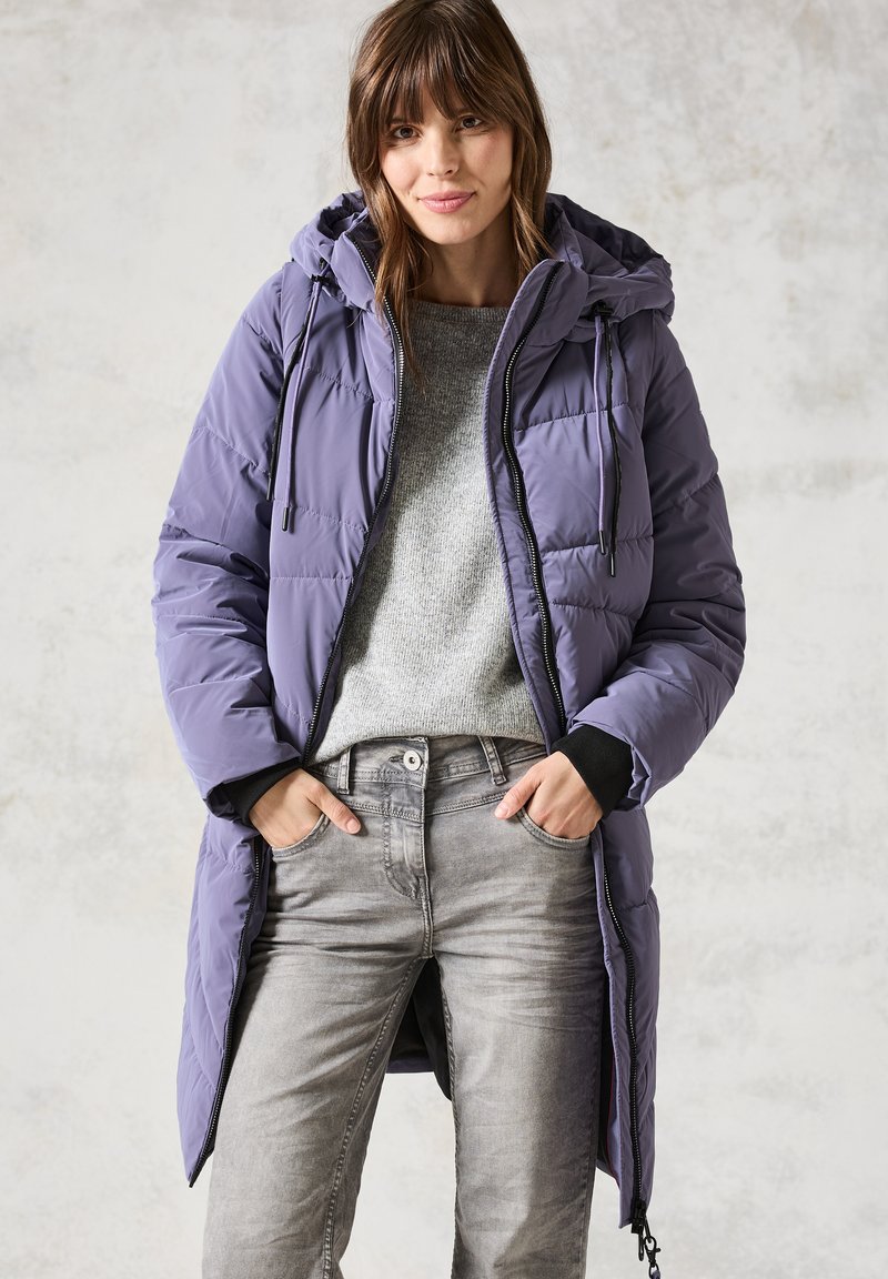 Abrigo largo acolchado morado con capucha, cierre frontal y cordones. Lleva puesto un suéter gris y jeans grises. Textura suave, brillo sutil.